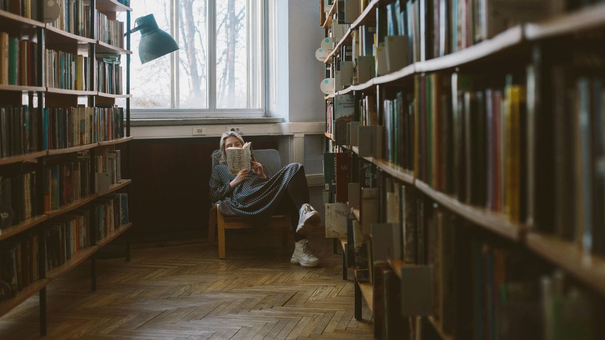 A relaxed woman sitting on a chair in a library and reading a book.