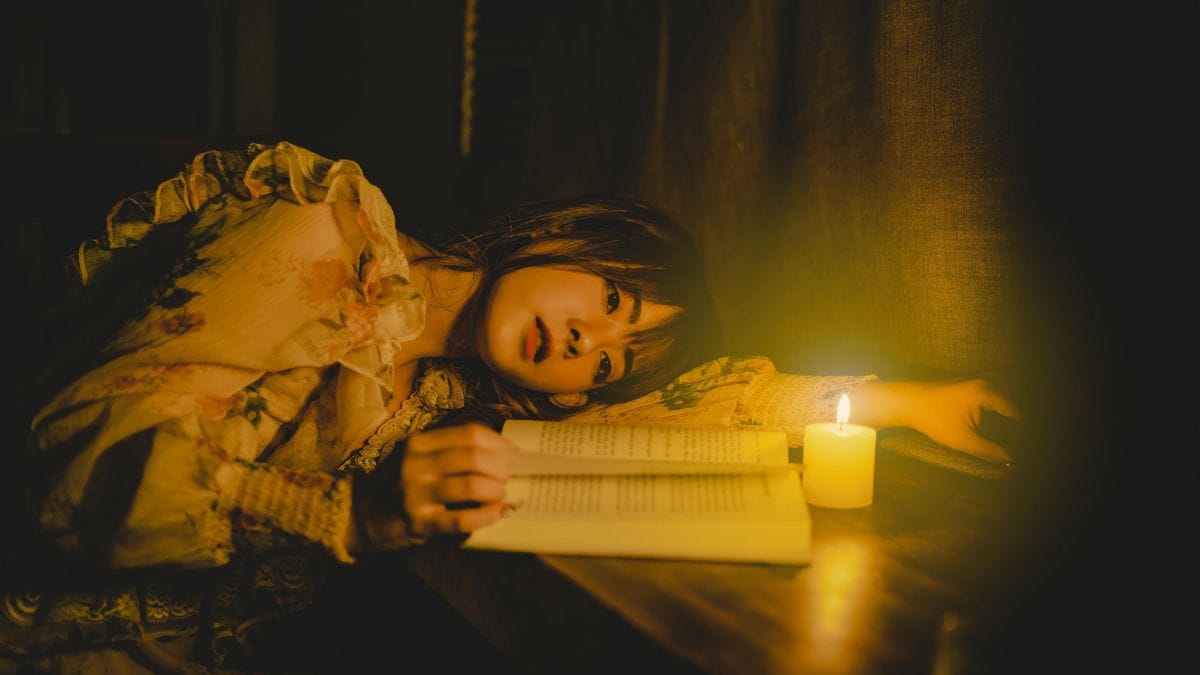 woman lying on a table with a wax candle beside her while reading a book