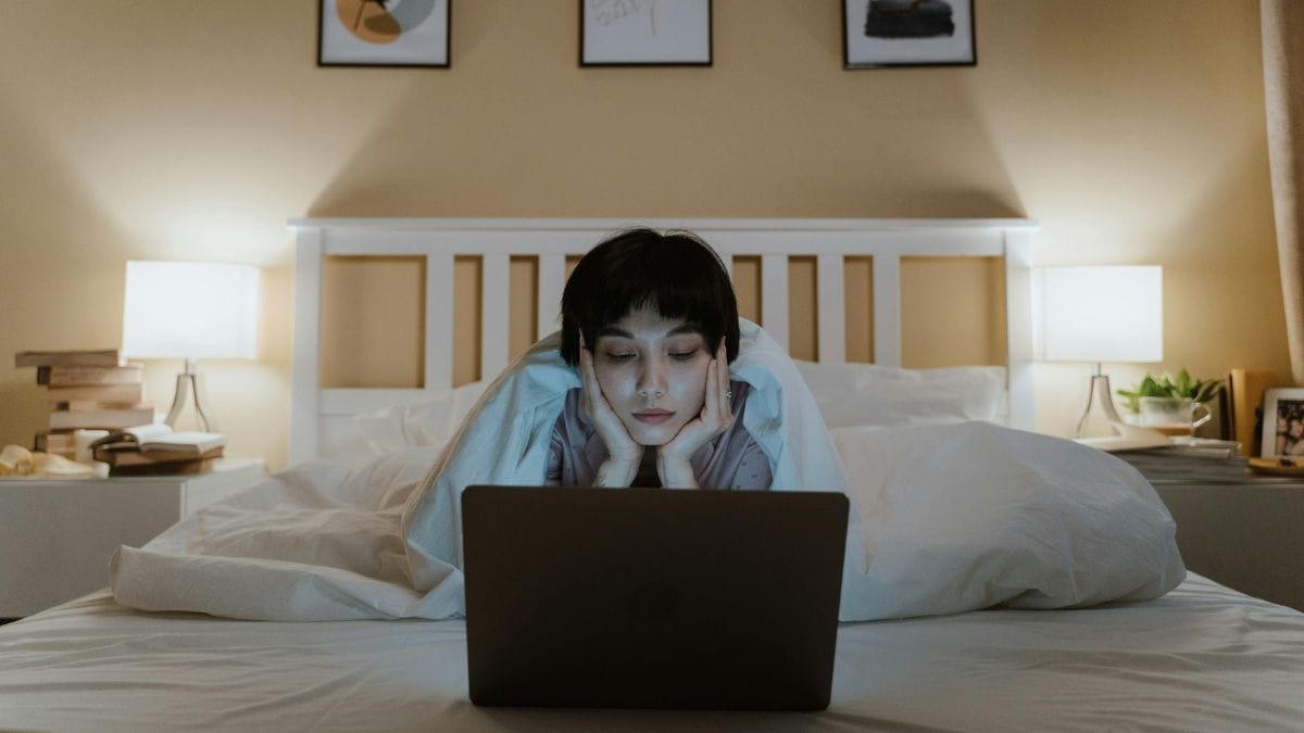 Person using laptop in bed at night with cozy bedroom lighting.