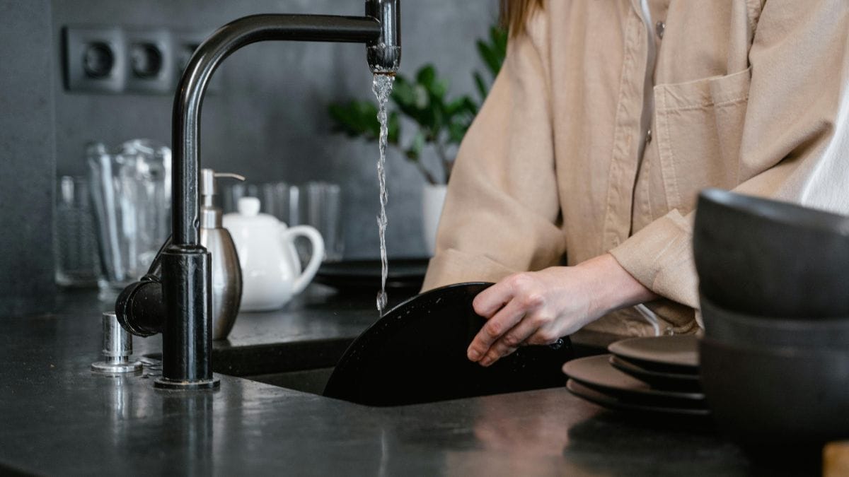 Person washing dishes at modern black kitchen sink with running water.