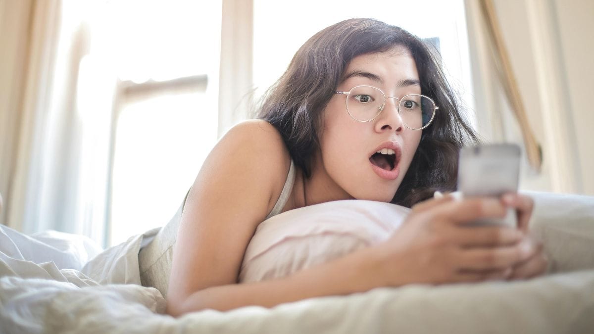 a woman lying on a bed, holding a phone with a shocked expression
