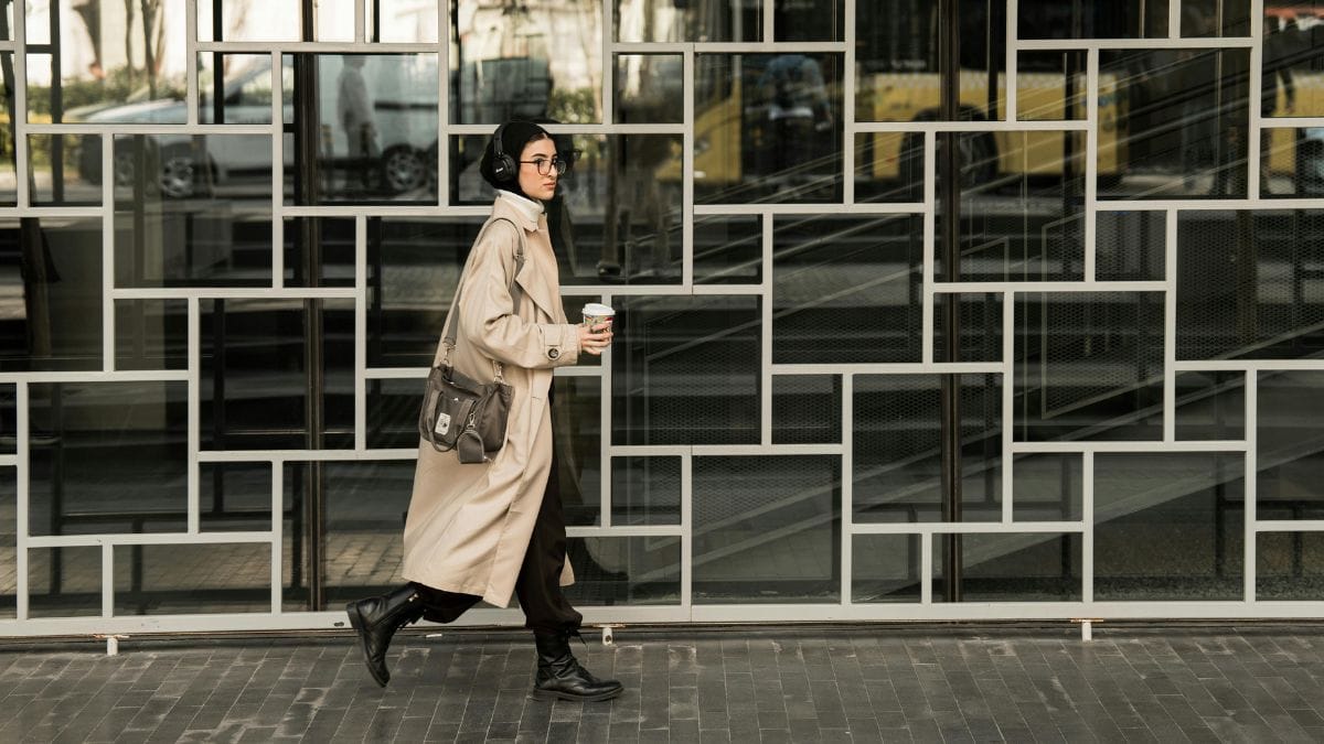 A stylish person walking through a modern city space, wearing headphones and holding a coffee, detached from the surroundings.