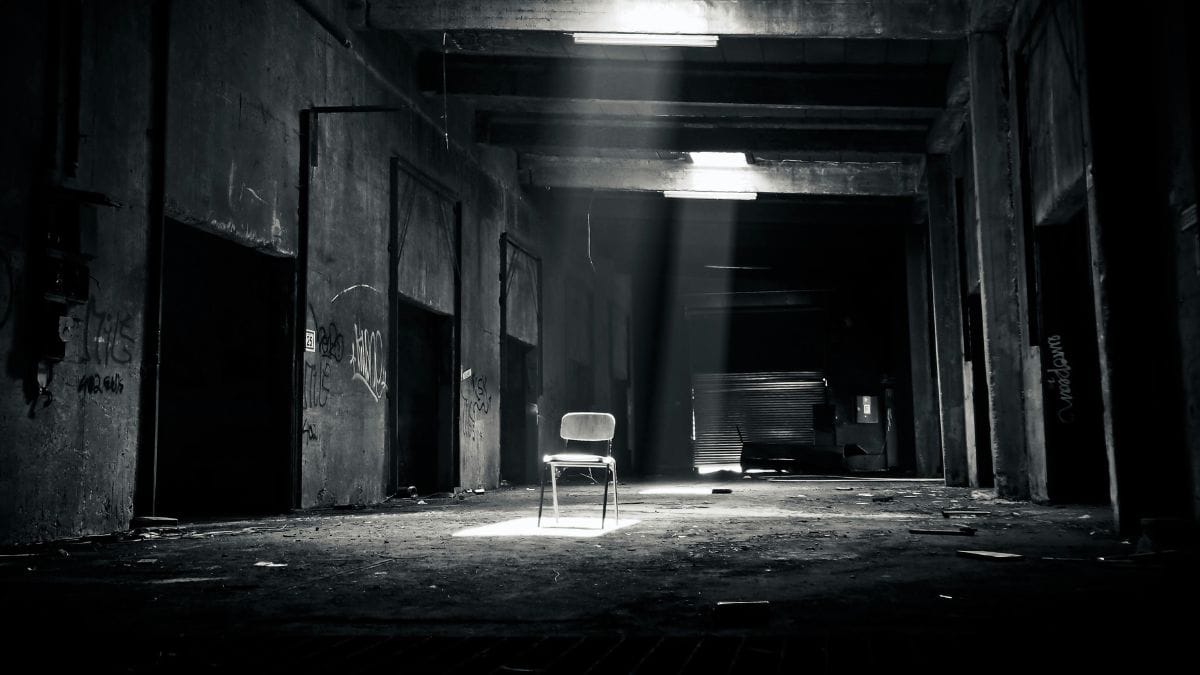 An empty chair illuminated by a spotlight inside a dark, abandoned industrial space.
