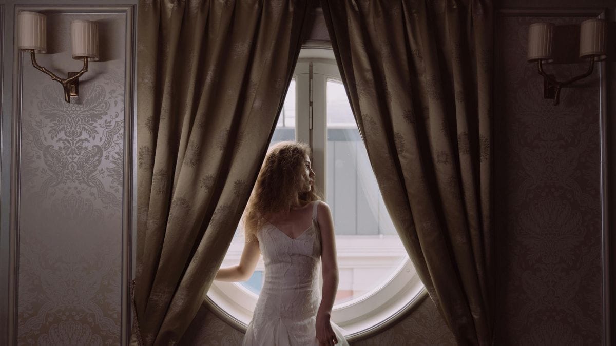 A woman in a white dress standing by a round window framed by heavy curtains in an elegant interior.